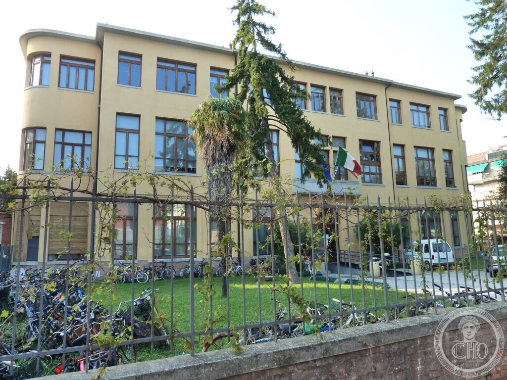 A three-storey yellow school building with a rounded corner, long rows of large windows, flags over the entrance, a palm and a cedar in the garden and dozens of bicycles against the iron fence