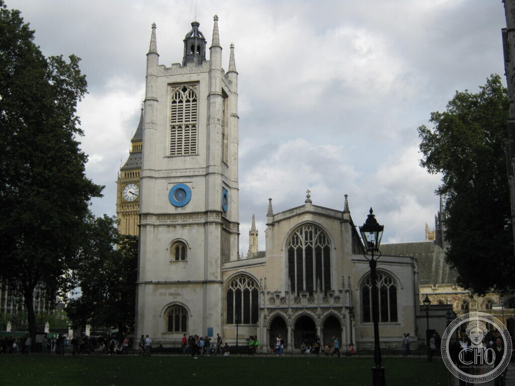 A pale stone Gothic church with a square west tower carrying two blue clock faces, the clock tower of the Houses of Parliament rising behind it on the left