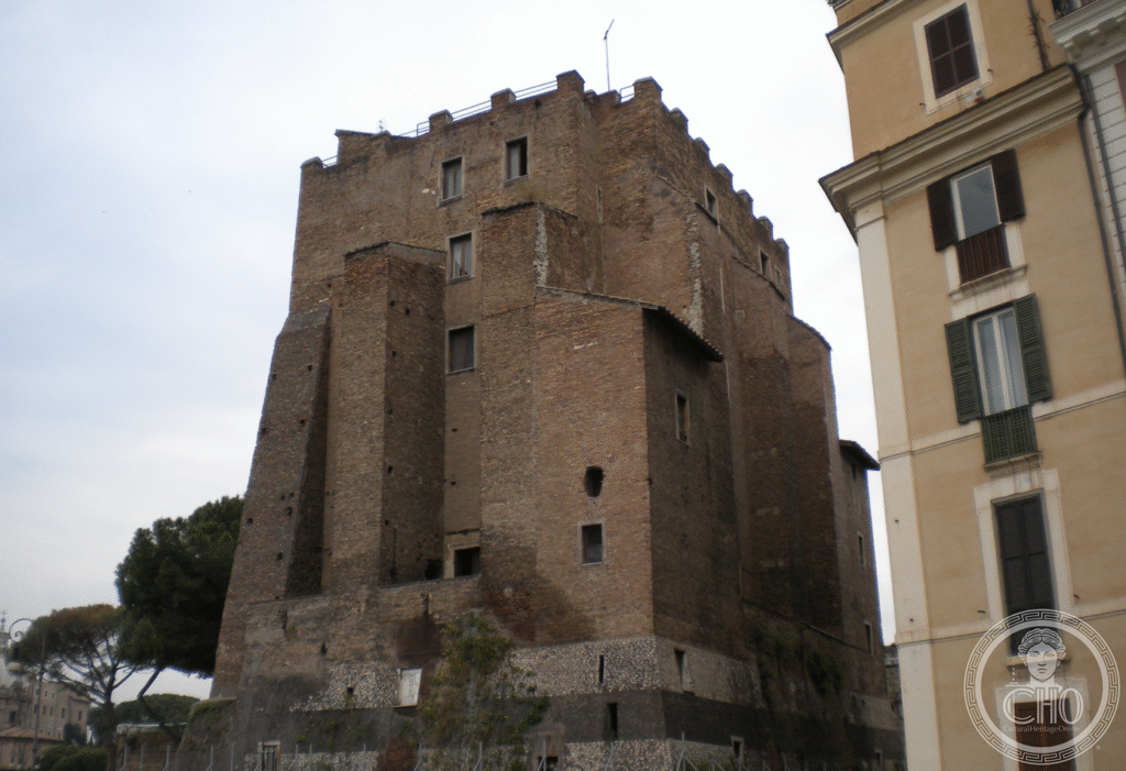 A massive truncated brick tower with sloping buttresses and a crenellated top, rising above a stone retaining wall beside a yellow palazzo