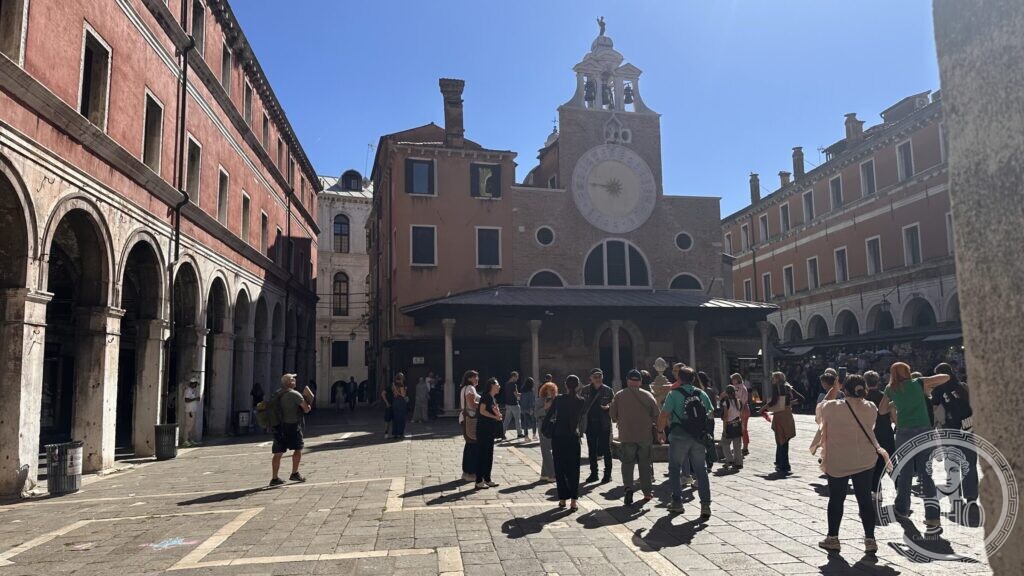 A group of walkers in a sunny square with arcades on the left and a small church with a large clock face at the end
