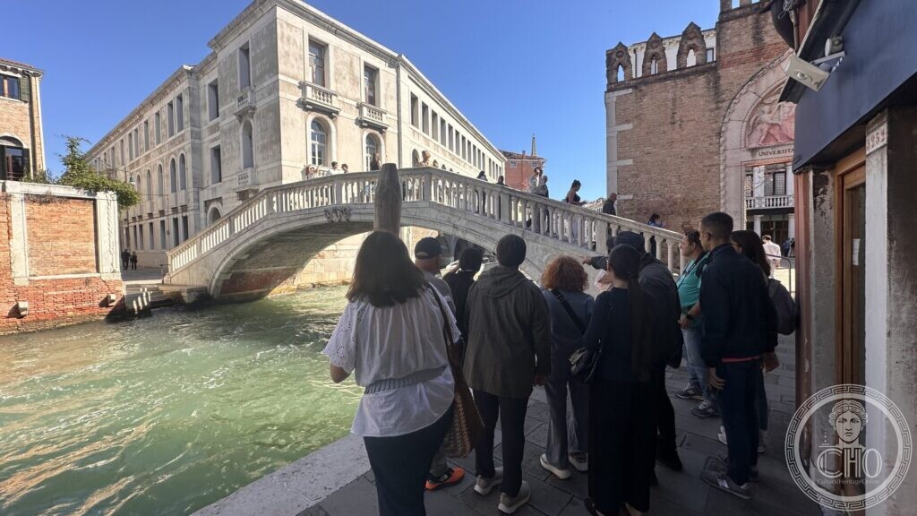 A group of walkers on a fondamenta looking at a white stone bridge over a green canal, with a pale palazzo and a brick gateway behind