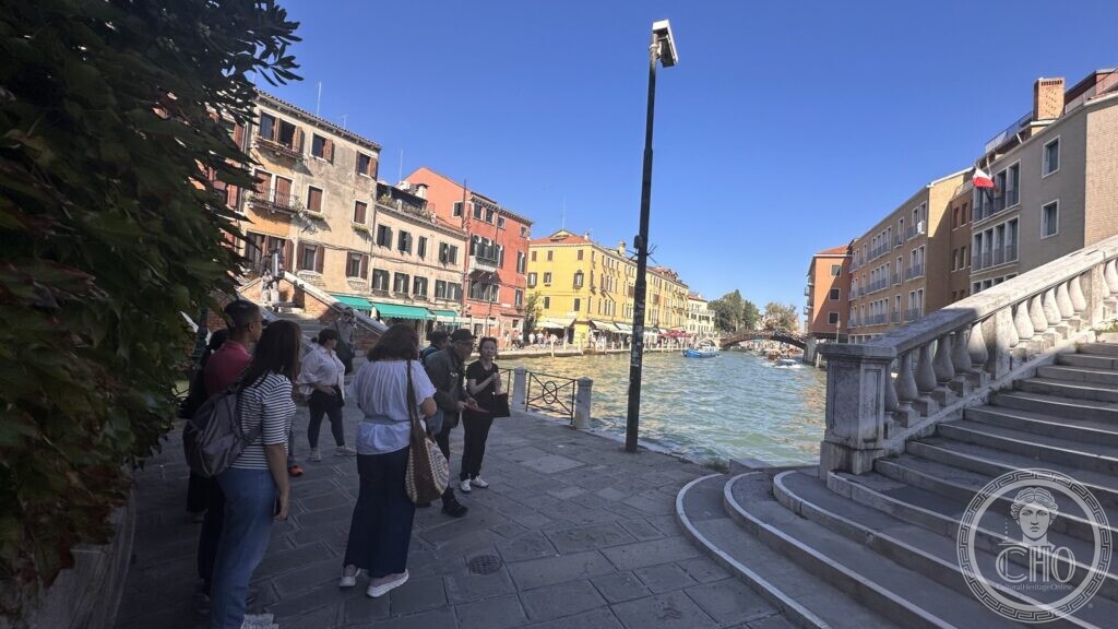 A group of walkers on a canal-side pavement beside the stone steps and balustrade of a bridge, coloured houses along the canal
