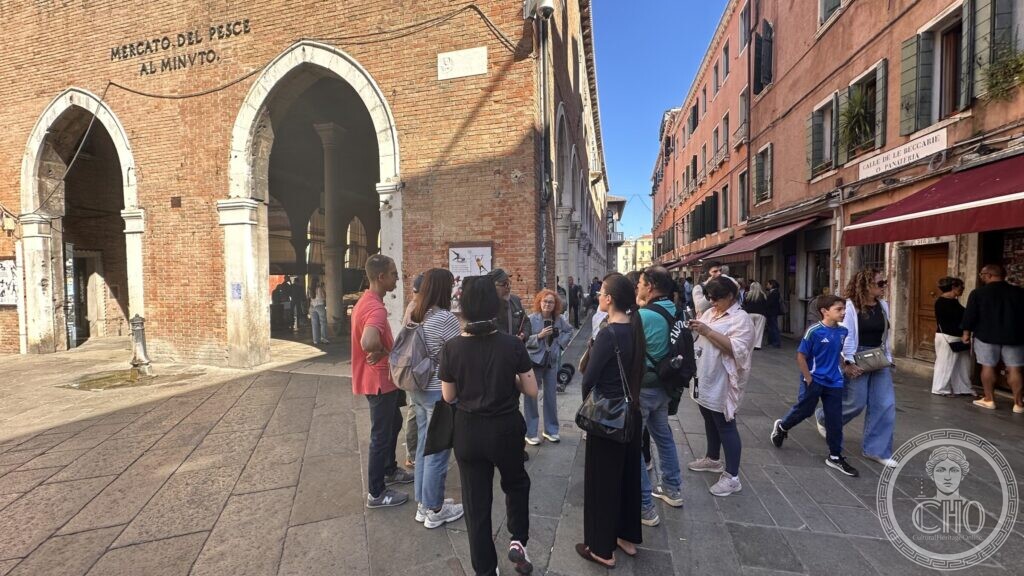A crowd of walkers in a paved square in front of a brick building with two pointed arches and the sign Mercato del Pesce al Minuto