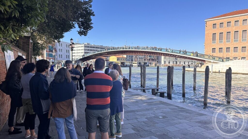A group of walkers on a wide canal-side pavement, wooden mooring posts in the water and a long modern bridge across the Grand Canal behind