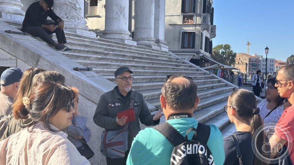 Walkers gathered at the foot of the columned steps of a church, a man speaking to them with a red notebook in his hand