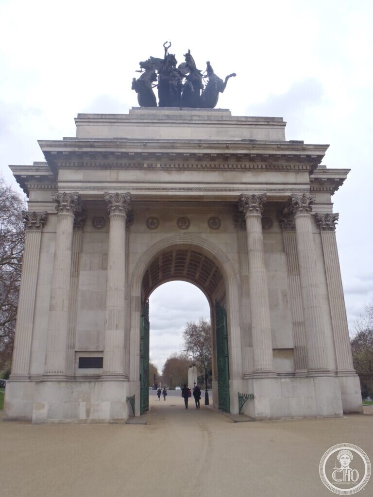 A Portland stone triumphal arch with Corinthian columns and green iron gates, crowned by a dark bronze group of four horses drawing a chariot