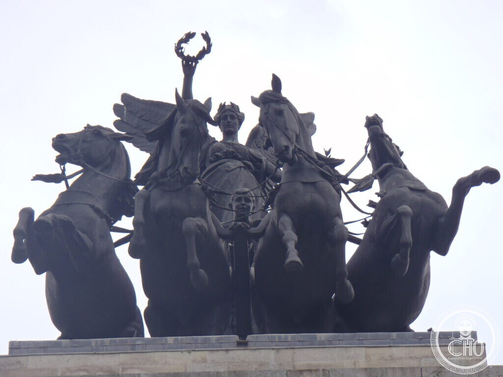 A bronze group of four rearing horses drawing a chariot, a winged female figure stepping down on to it holding out a laurel wreath, seen against a pale sky