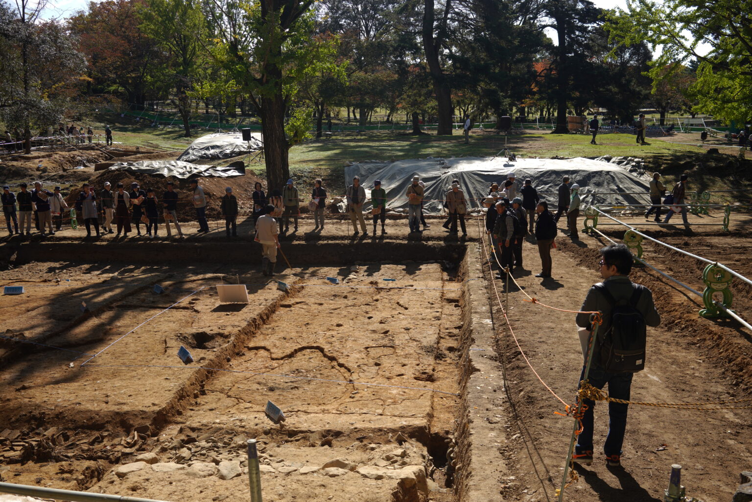 Archaeologists and visitors standing around a roped excavation trench cut into bare ground inside the Tōdai-ji precinct