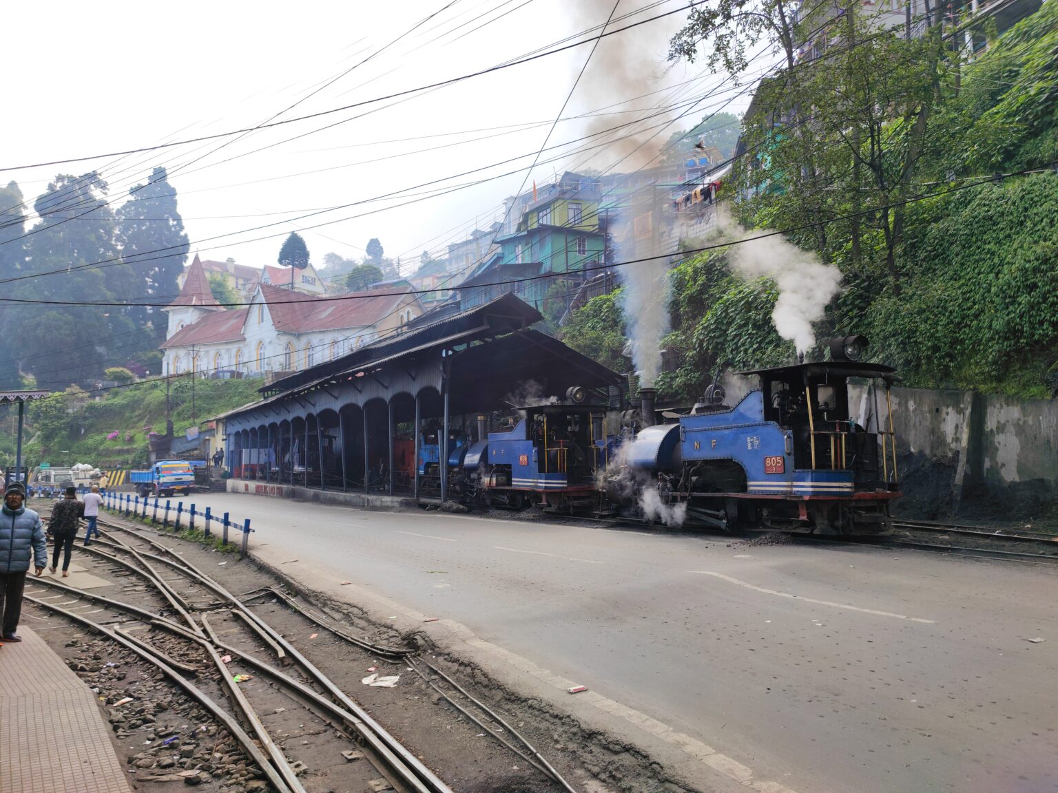 A small steam locomotive standing under its own smoke beside a station platform, with the town on the slope behind