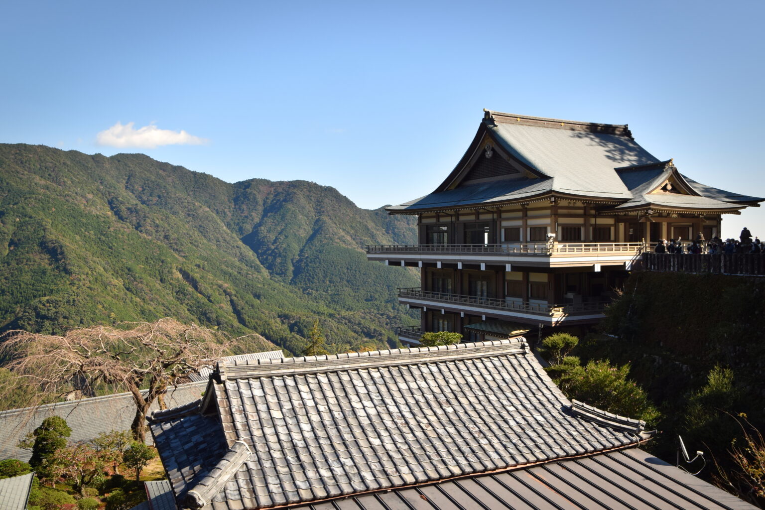 A tall temple hall with tiled roofs and a viewing gallery, standing above wooded mountain ridges at Nachi