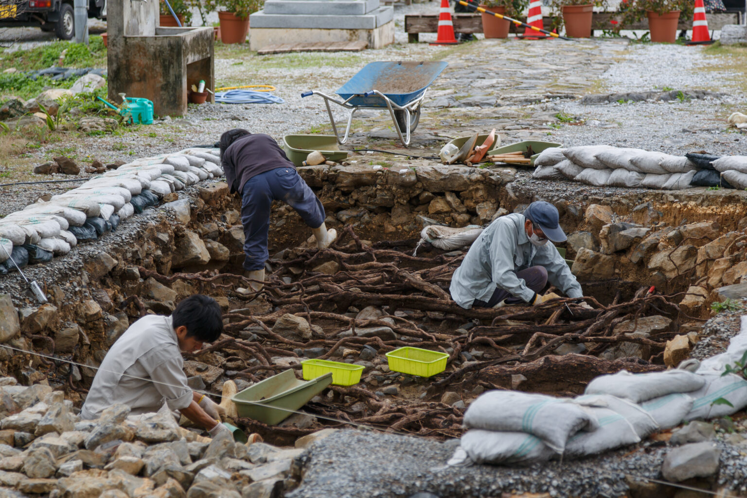 Archaeologists working in an open excavation trench in front of a stone wall, with tools and sandbags